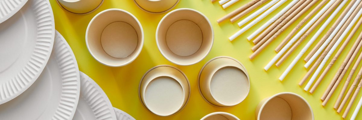 cardboard straws in the upper right corner a row of cardboard cups in the center and some white cardboard plates in the lower left corner of the image on a yellow background